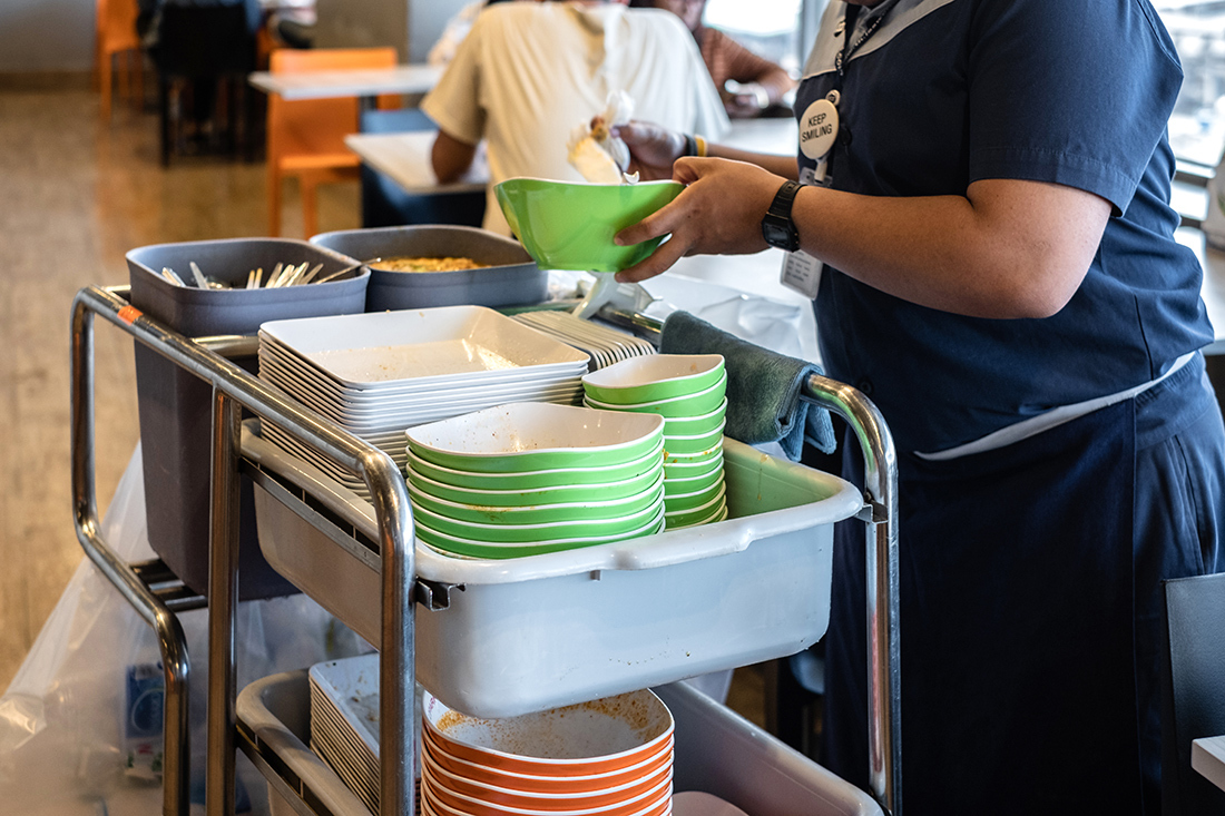 Cafeteria service worker collects dirty dishes and places them in a bus tub on a meal cart